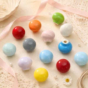 Pode incluir: Uma coleção de puxadores de armário em cerâmica, redondos e lisos, em várias cores sólidas, incluindo vermelho, laranja, amarelo, verde, azul, cinzento, branco e tons pastel. Cada puxador tem um acabamento brilhante e um orifício central.