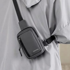 May include: A dark gray crossbody sling bag with a black adjustable strap and a front zipper pocket featuring the word FASHION printed in white. The bag is worn over a light gray hooded jacket and dark gray trousers, showcasing a compact design.