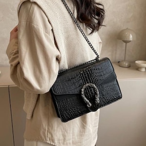 Puede incluir: Un bolso de hombro rectangular negro con un acabado texturizado de piel de cocodrilo, que cuenta con una correa de cadena metálica plateada y un prominente cierre plateado en forma de herradura con detalles de cabeza de animal.