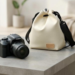 May include: A black Canon DSLR camera with a lens attached sits next to a cream-colored drawstring bag. The bag features a black strap and a small, rectangular tan leather-colored patch on the front with the embossed letters XBY.