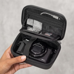 May include: A black, rectangular, zipped hard-shell carrying case containing a black Canon G7X digital camera secured by an elastic strap. The interior lid features a zipped mesh pocket and a secondary mesh compartment for small accessories.
