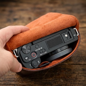 Puede incluir: Una cámara digital negra con lente y controles superiores, incluyendo un botón de disparo con anillo rojo e interruptor de encendido, parcialmente dentro de una funda de cuero ante color canela con forro suave y herrajes metálicos.