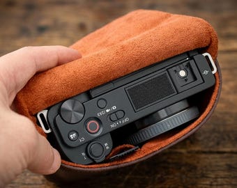 May include: A black digital camera with a top-mounted shutter button, a red-ringed record button, and a mode switch, partially enclosed in a brown leather protective case with a soft, textured interior lining.