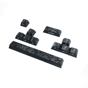 May include: A set of black and grey forged carbon fibre keyboard keycaps featuring a marbled pattern. The collection includes an Esc key, W, A, S, D keys, an Enter key, a long spacebar, and four arrow keys, all with white printed lettering.
