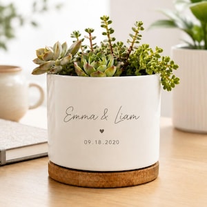 Puede incluir: Una maceta cilíndrica de cerámica blanca sobre una base circular de madera. La maceta presenta el texto impreso "Emma & Liam" sobre un pequeño icono de corazón y la fecha "09.18.2020". Contiene una variedad de plantas suculentas verdes.