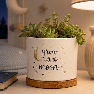 May include: A white cylindrical ceramic planter with a cork base, containing green succulents. The planter features a gold crescent moon, small gold stars, and the black text "grow with the moon" printed on the front, along with a constellation design.