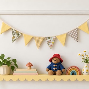 May include: A fabric bunting banner with yellow, gingham, and floral triangular flags hangs above a scalloped yellow shelf. The shelf holds a potted plant, a mushroom figurine, books, a teddy bear, a rainbow toy, a flower vase, and a house block.