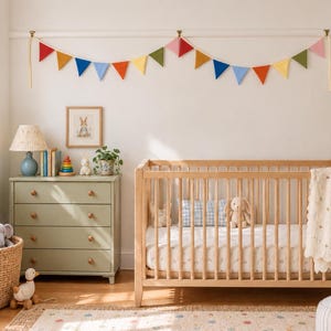 Puede incluir: Una cuna de madera clara con barrotes verticales, un colchón con estampado blanco y una manta color crema con borde festoneado. Sobre la cuna cuelga una guirnalda de tela colorida con banderines triangulares en tonos rojo, naranja, amarillo, verde, azul y rosa.