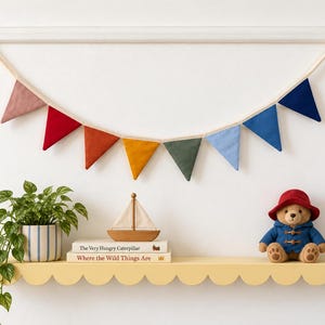 Op de afbeelding: Een stoffen vlaggenlijn met acht driehoekige vlaggetjes in gedempte tinten roze, rood, oranje, geel, groen, lichtblauw, blauw en donkerblauw, opgehangen aan een crèmekleurig koord bevestigd aan twee goudkleurige muurhaken.