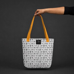 May include: A white tote bag featuring a repeating black geometric pattern of lines, circles, and crosses. The bag has two mustard yellow shoulder straps and a small black rectangular label in the center with the white letters ND printed on it.