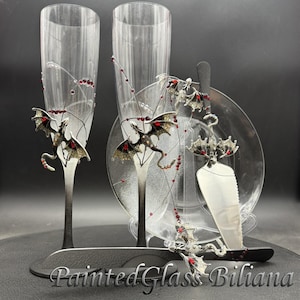May include: Two clear glass champagne flutes with black ombre stems, a matching cake server, and a knife. Each piece features a silver-toned dragon figurine with red crystal accents and delicate wire detailing. The text PaintedGlassBiliana is visible.