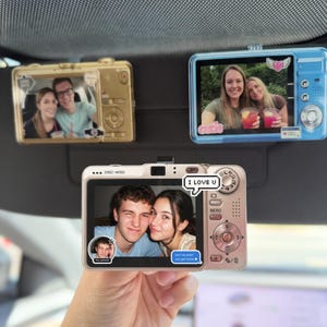 May include: Three digital camera-shaped car visor clips featuring photo displays. The gold clip shows a couple, the blue clip shows two people with drinks, and the rose gold DSC-W50 clip displays a couple with text bubbles saying I LOVE U and text me when you get home.