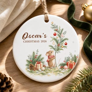 May include: A round white ceramic ornament featuring a brown mouse wearing a red scarf, standing next to a decorated Christmas tree with red and gold ornaments. The text reads Oscar's CHRISTMAS 2026. It hangs from a jute twine loop.