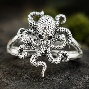 Op de afbeelding: Een zilverkleurige ring met een gedetailleerd octopusontwerp. De octopus heeft een textuur met stippen op de kop en tentakels, waarbij donkere, verdiepte delen de zuignappen en ogen accentueren. De ringband is glad en gepolijst.