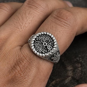 May include: A silver-toned ring featuring a circular face with a textured, dotted background and a raised chemical structure symbol. The ring band is framed by a twisted rope-like border and includes a small leaf emblem on the side of the shank.