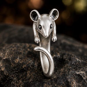 Puede incluir: Un anillo de tono plateado con forma de ratón, que presenta una cabeza detallada con orejas prominentes y ojos oscuros, con el cuerpo formando la banda y la cola curvándose hacia arriba para completar el diseño circular de la pieza.