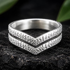 Pode incluir: Um anel de prata em forma de chevron com uma superfície texturizada e martelada em toda a banda exterior. O anel tem uma ponta em forma de V no centro, criando um design simétrico com uma banda interior polida e um acabamento exterior rugoso.