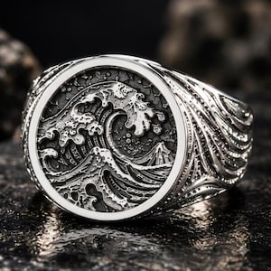 May include: A silver-toned metal ring featuring a circular face with a detailed, raised relief design of a crashing ocean wave and a mountain peak. The band of the ring is textured with flowing, wave-like patterns that wrap around the sides.