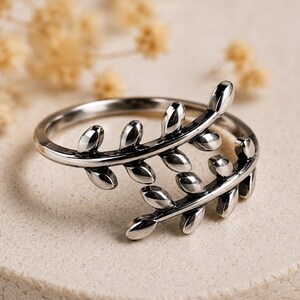 May include: A polished silver-toned ring featuring an open-ended design that resembles two overlapping leafy branches. The band is crafted with multiple small, smooth, oval-shaped leaf motifs extending along the curved metal structure.