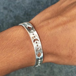 May include: A silver-toned cuff bracelet featuring a series of cut-out celestial shapes, including stars, a crescent moon, and various circular dots arranged in a linear pattern along the centre of the polished metal band.