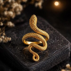 May include: A gold-toned ring shaped like a coiled snake with a textured, scale-like surface. The snake's head is positioned at the top, while its body wraps around to form the band, ending in a curled tail at the bottom of the design.