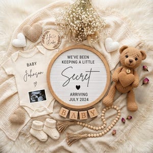 Könnte beinhalten: Ein cremefarbener Babybody mit dem Text Baby Johnson und einem Ultraschallbild, ein rundes Buchstabenbrett mit der Aufschrift We've been keeping a little secret arriving July 2024, ein gehäkelter Teddybär, Holzwürfel und kleine Strickherzen.