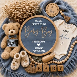 May include: A round blue felt letter board with the text WE ARE EXCITED TO SAY Baby Boy IS ON THE WAY, a white heart, and ARRIVING AUGUST 2026. Includes a tan teddy bear, cream baby bodysuit with Baby Adam, ultrasound photo, booties, and wooden blocks.