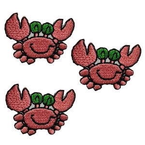 Könnte beinhalten: Drei gestickte Aufbügler, die jeweils ein stilisiertes Krabbenmotiv zeigen. Die Krabben sind überwiegend rosa mit schwarzen Stickkonturen, haben zwei leuchtend grüne Augen auf Stielen sowie große Scheren und sind in Dreiecksform angeordnet.