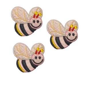 May include: Three embroidered bee patches featuring a light pink face and body with black and yellow stripes. Each bee has two light pink wings with gold stitching, red antennae with yellow tips, and a stitched black smiling mouth and eyes.