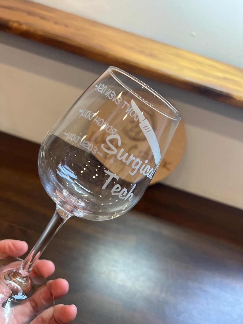 May include: A clear glass wine glass with the text "Surgical Tech" and a small illustration of a scalpel etched into the side.