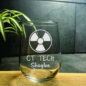 May include: A clear glass wine glass with a white radiation symbol and the text "CT TECH Shaylee" etched on the side.