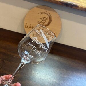May include: A clear glass wine glass with the text "Surgical Teeth" etched on the side. The glass is held in a hand.