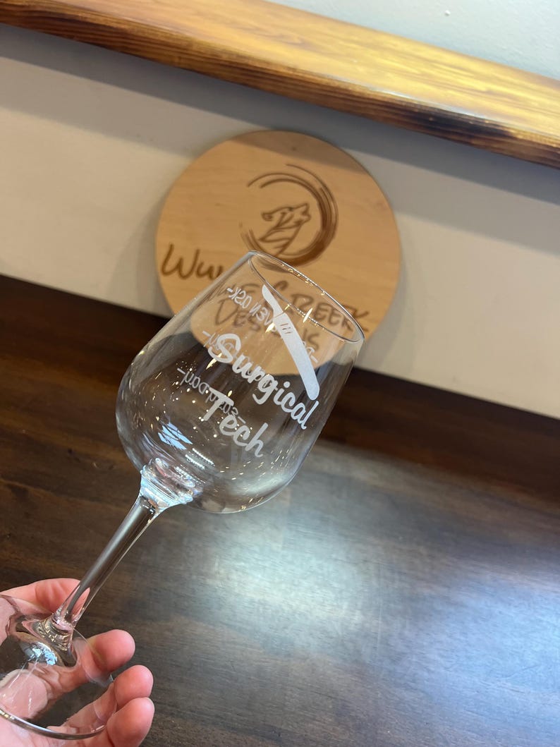 May include: A clear glass wine glass with the text "Surgical Teeth" etched on the side. The glass is held in a hand.
