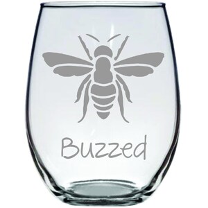 May include: A clear glass wine glass with a gray bee silhouette and the word "Buzzed" etched on the bottom.