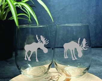 May include: Two clear glass wine glasses with etched moose silhouettes. The moose are facing each other and are in a light gray color.