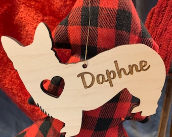 May include: A wooden corgi-shaped ornament with the name "Daphne" engraved. It features a heart-shaped cutout and hangs in front of a red and black check fabric Christmas tree. The background is a blurred red.