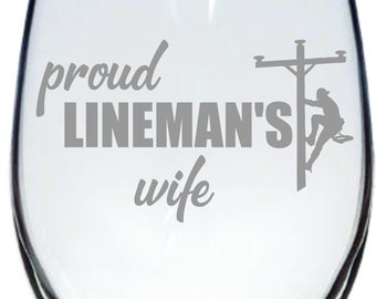 May include: A clear glass wine glass with the text "proud lineman's wife" and a silhouette of a lineman working on a power line.