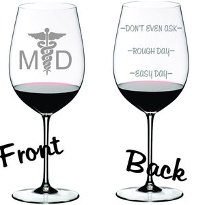 May include: A clear glass wine glass with a gray medical symbol and the letters "MD" on the front. The back of the glass has the text "Don't even ask", "Rough day", and "Easy day" written on it.