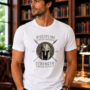 Può includere: Una t-shirt bianca a maniche corte con una stampa grafica di un elmo spartano centrato sopra due lance incrociate e uno scudo circolare. Il testo recita DISCIPLINE BUILDS FREEDOM, EST. 480 BC e STRENGTH BUILDS DESTINY in grigio scuro.