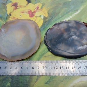 Può includere: Due fette circolari di agata posizionate sopra un righello di metallo. La fetta a sinistra è beige e bianca traslucida, mentre quella a destra è grigio scuro e nera con una superficie lucida. Il righello indica una larghezza di 9 cm.