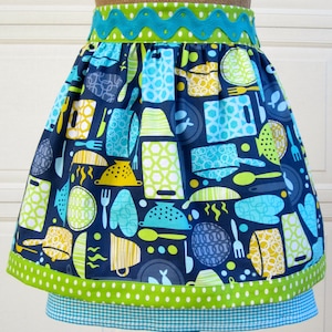 May include: A blue and green apron with a kitchen-themed print. The apron has a green and white polka dot trim and a blue and white gingham trim.