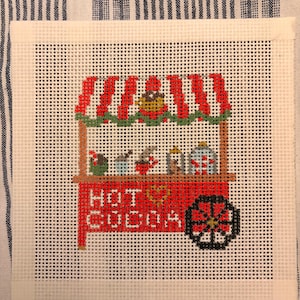 May include: A needlepoint canvas featuring a red and white striped hot cocoa stand with a green garland. The stand displays mugs and jars, with the text "HOT COCOA" and a heart symbol stitched in red on the front. A black wheel is on the right side.