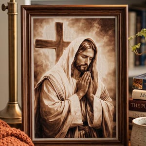 May include: A framed sepia-toned print depicting a figure with long hair and a beard, hands clasped in prayer, positioned before a wooden cross. The artwork is displayed on a wooden table next to a stack of books, a ceramic mug, and a lamp.