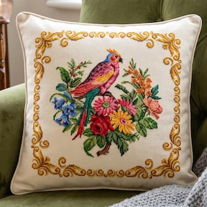 May include: A square off-white throw pillow featuring a cross-stitched design of a colorful parrot perched among vibrant flowers. The central motif is surrounded by a decorative gold-colored border, creating a detailed and ornate appearance.