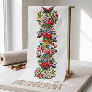 May include: A white rectangular linen runner featuring a vertical embroidered design of three colorful parrots perched among vibrant red, pink, and purple flowers with green foliage. The runner has a fringed bottom edge and hangs on a gold stand.