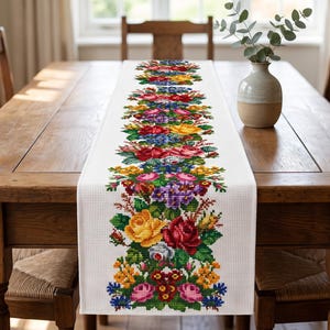 May include: A white rectangular table runner featuring a vibrant, repeating cross-stitch floral pattern. The design displays a dense arrangement of roses and various small blossoms in shades of red, yellow, purple, blue, and green.