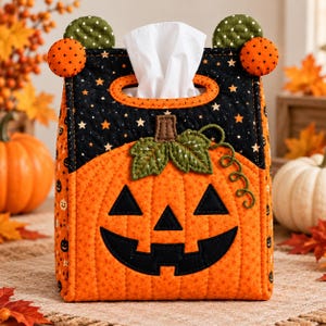 May include: A quilted fabric tissue box cover featuring a jack-o'-lantern face on an orange background with a black star-patterned top. The cover includes green fabric leaves, a curly vine, and four small circular accents at the top corners.