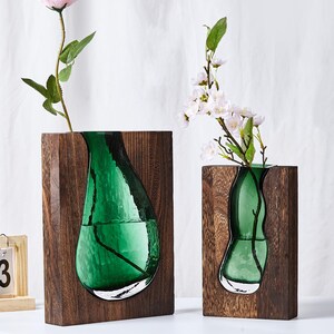 May include: Two rectangular dark brown wooden blocks each featuring a recessed, organic-shaped green glass vase insert. One vase holds a single pink rose, while the other holds a flowering branch. A small wooden calendar shows the text DEC 13.