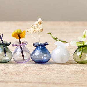 May include: A set of six miniature glass vases in various colours including dark green, light purple, deep blue, opaque white, olive green, and black. Each small, rounded vessel features a ruffled, flared rim and holds a single small stem or flower.