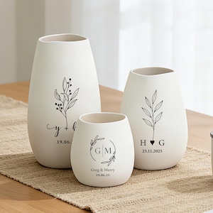 Op de afbeelding: Drie matwitte keramische vazen van verschillende hoogtes, elk voorzien van zwarte botanische lijntekeningen en gepersonaliseerde tekst. De ontwerpen bevatten de initialen Y+K met 19.06.25, G|M met Greg & Marry 19.06.25 en H hart G met 23.11.2025.